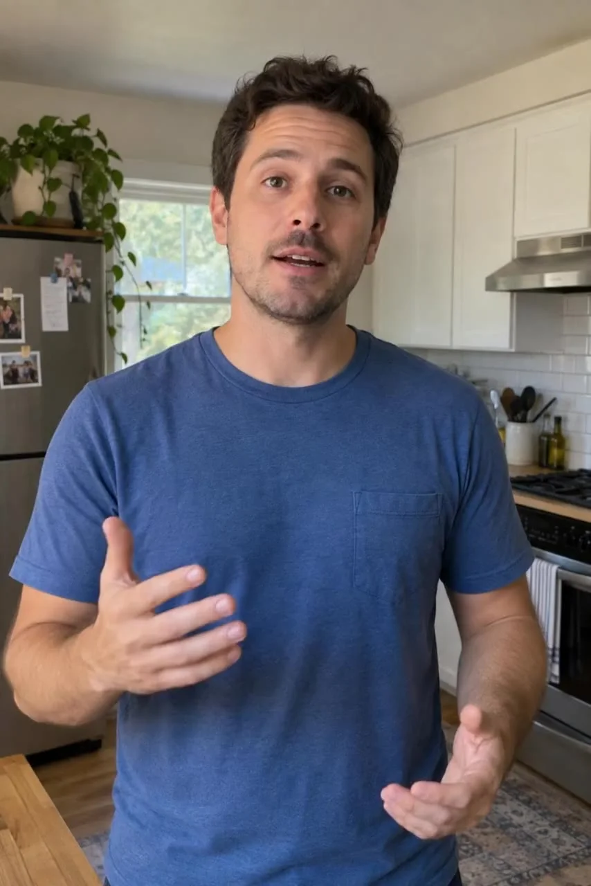 Starting frame: A man in a plain blue T-shirt explains something to the camera with one hand in a home kitchen.
